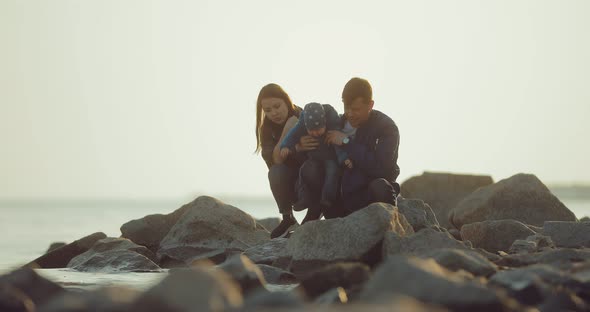 Husband Wife and Baby Sit on the Rocks on the Rocky Coast and Look Into the Distance alt