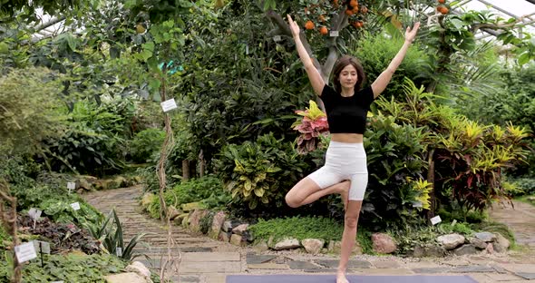 A Beautiful Brunette Woman is Doing Yoga Exercises on a Mat in a Beautiful Botanical Garden
