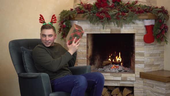A Young Man Sits in an Armchair By the Fireplace with Horns on His Head and Holds and Shakes Gifts alt