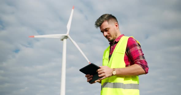 The Engineer Work Next to the Windmill Turbines with the Tablet in His Hand alt