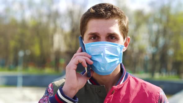 Young guy in medical mask talking on mobile phone in park alt