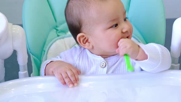 A hungry baby is gnawing on a plastic spoon at the table on a high chair. Teething, whims, itchy gum alt
