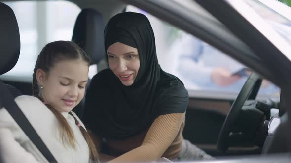 Positive Smiling Mother in Hijab Fastening Seat Belt for Daughter Sitting in Car alt
