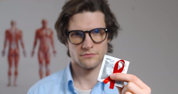 Close Up Portrait of Young Man in Glasses Holding Condom with Aids Awareness Red Ribbon alt