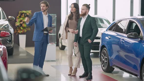 Wide Shot Portrait of Confident Young Rich Couple Choosing New Automobile in Car Dealership alt