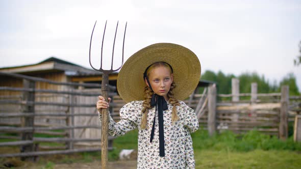 Serious Girl in Dress and Wicker Hat Posing with Hayfork in Village alt