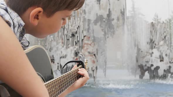Caucasoid Teenager in a Plaid Shirt Plays an Acoustic Guitar While Sitting By a Water Fountain alt