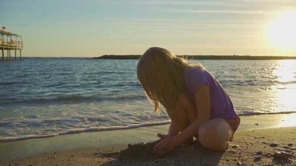 a Cute Little Girl Sits on the Seashore Playing in the Sand and with Pebbles alt