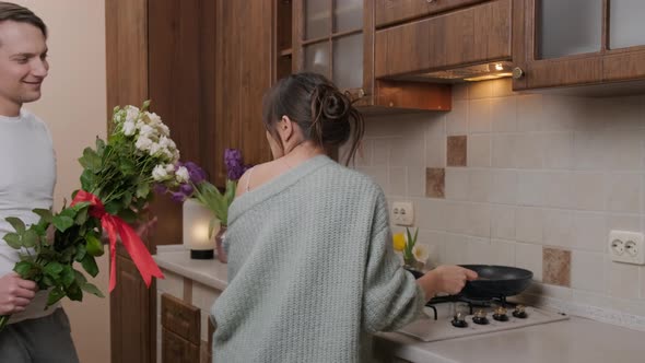 Young Man Giving Flowers To His Wife Who Prepares Food in the Kitchen alt