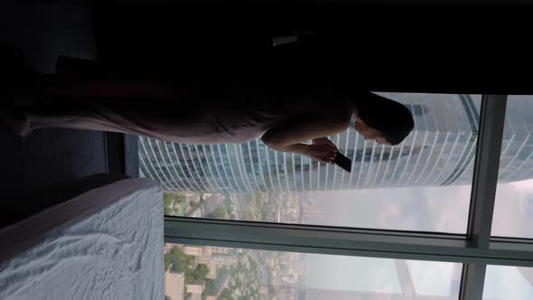 Woman in a Nightgown Approaches a Panoramic Window in a Skyscraper alt