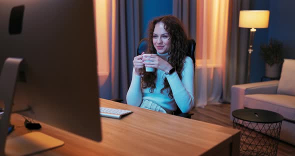Tired After Work Pretty Girl Relaxes in Evening Sitting in Front of a Computer Screen alt