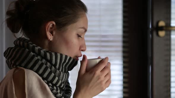 A sick woman warms up and drinks a hot drink near the window on a winter evening alt