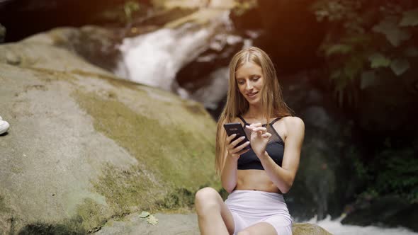 A Smiling Young Woman is Using Smartphone and Typing Messages with Beautiful Nature Background alt