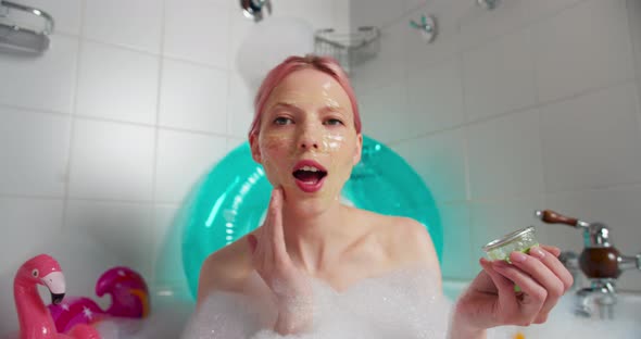 Cheerful Woman with Pink Hair Sits in a Bath in a Cloud of Foam and Makes a Cosmetic Mask alt