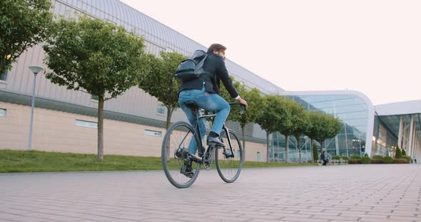 Guy Riding a Bicycle to His Work on Cobbled Path Near Modern Big Bulding with Glass Windows alt