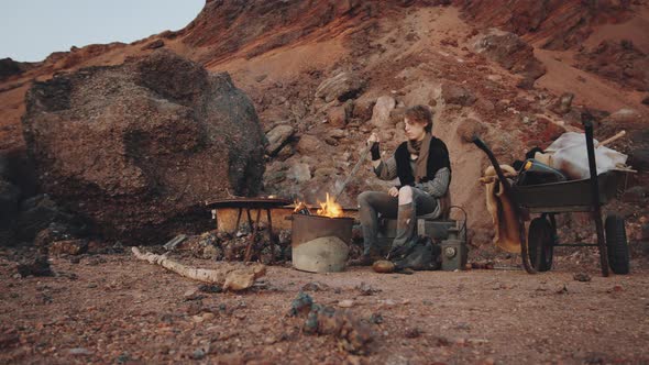 Woman Stirring Embers in Campfire in Post Apocalyptic World alt