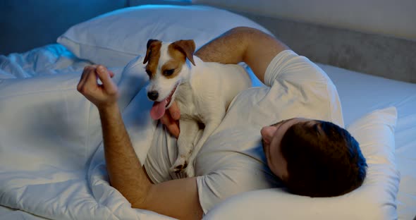 a Man Is Lying on a Sofa with White Bed Linen with His Dog. the Guy Is Stroking the Animal's Neck alt