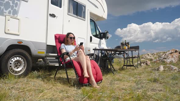 Woman Sitting Near Camper Rv On Folding Chair Using Sell Phone On Camp Site alt