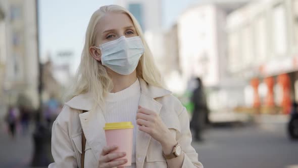 A Young Woman in a Medical Mask Stands with Coffee in Her Hands on a Central Street and Waits for