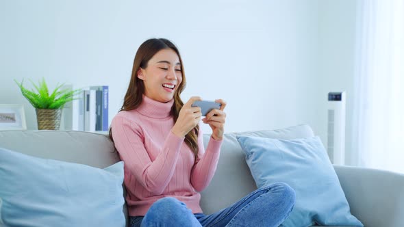 Asian young beautiful woman playing mobile game on smartphone at home feel happy and relax. alt