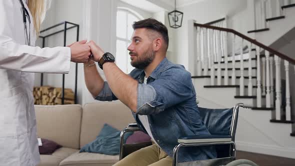 Medical Worker Helping Handsome Confident Positive Young Bearded Man Get Out from Wheelchair alt