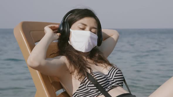 Young Woman in Medical Mask Relaxing on Beach Deck Chair By the Sea with Eyes Closed Wearing alt