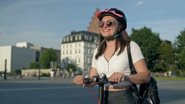 Young Cheerful Woman in Sunglasses and a Helmet Stands on an Electric Scooter in the Middle of a