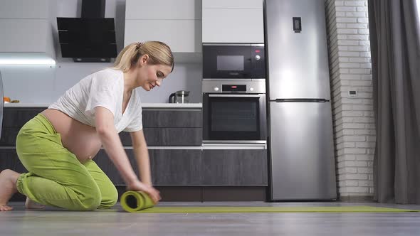 A Young Pregnant Woman Folds a Green Yoga or Fitness Mat After a Workout at Home in the Living Room