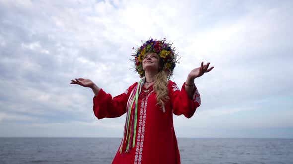 Smiling Ukrainian Woman in Red Dress and Head Wreath Raising Hands Looking Up at Cloudy Sky alt