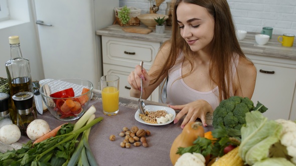 Lovely Girl Eating Raw Sprouts Buckwheat with Nuts in Kitchen with Fresh Vegetables and Fruits alt