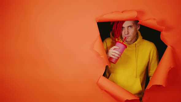 Young Man Drinking Through Soft Drink Straw in Hole of Orange Background alt