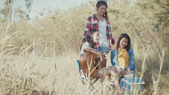 Three young adult friends Asian women happy camping in nature having fun together playing guitar.