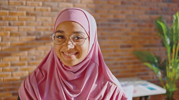 Muslim Woman in Pink Hijab and Glasses Smiling and Looking at the Camera Against the Background of a alt