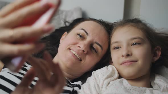 A Happy Woman and Her Daughter Lie Together on a Bed Take Selfies on a Smartphone or Communicate Via alt