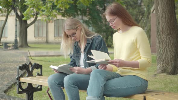 Two Concentrated Nerd Female Students in Eyeglasses Sitting on Bench and Reading Books. Portrait of alt