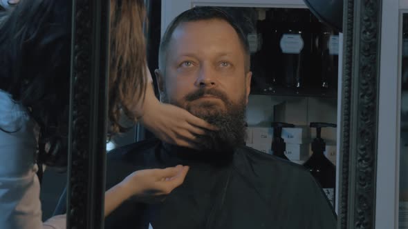 Close up view of female barber taking cares of lush beard of man Barber applying cosmetics alt