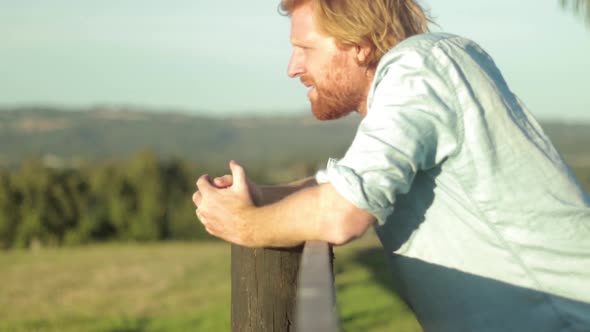 A young farmer in a blue shirt rests on a wooden fence post and looks out across the country in slow alt