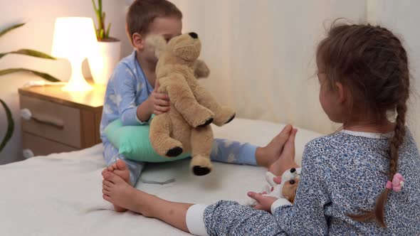Two Smiling Preschool Toddler Children In Pajamas Playing With Teddy Bear on Bed
