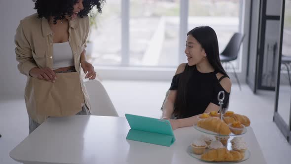 African American Young Woman Entering Living Room Bringing Takeaway Food for Asian Friend Working in alt