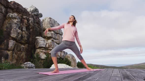 Caucasian woman practicing yoga outdoors, stretching standing on deck in rural mountainside setting alt