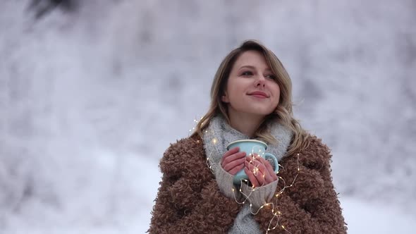 Beautiful girl in coat with cup of hot drink in a snow forest.