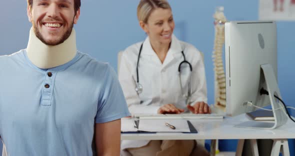 Patient smiling at camera while female physiotherapist working over computer in background alt
