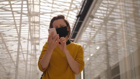 Busy Cheerful Girl with Protective Face Mask Walks Looking at Her Phone in in Shopping Center. Glass alt