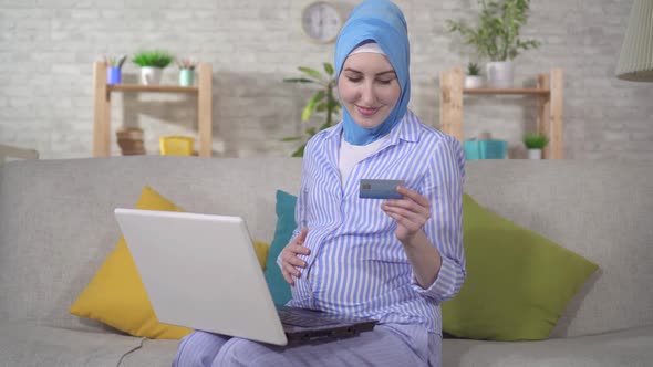 a Young Pregnant Muslim Woman in Hijab Uses Laptop and Bank Card alt