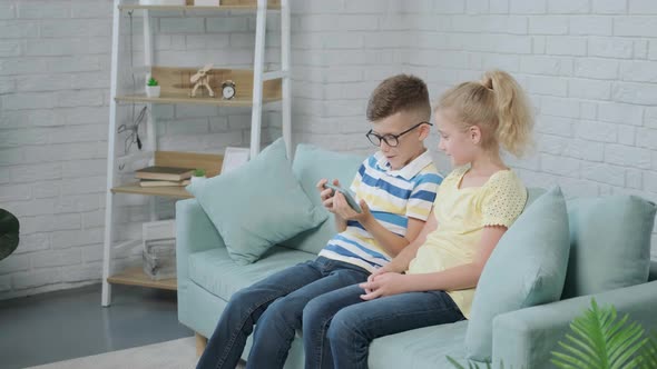 Excited Twins Kids Using Smartphone Sitting Together on Sofa alt