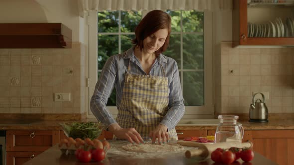30s Woman in Apron Placing Grated Cheese on Pizza Dough Standing in the Kitchen alt