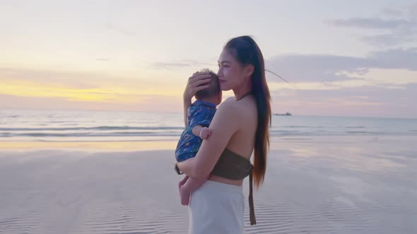 Happy mother with baby boy walks by ocean on the beach in summer alt
