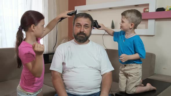 A young son and daughter cut their father's hair on his head with an electric clipper