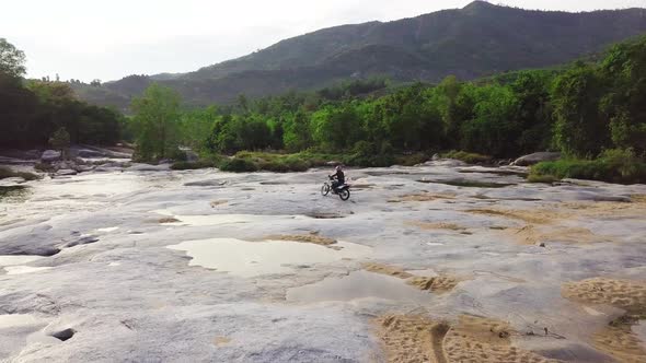 Aerial: Man Driving a Motorcycle on Paved Stone Surface with Sand in the River. alt