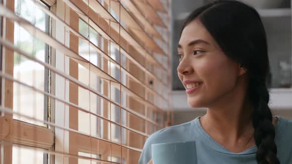 Asian woman smiling and drinking coffee beside a window at home.